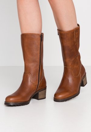 Mid-calf brown leather boots with side zippers, block heels, and textured soles worn on bare legs against a white background.