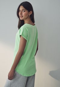 Hellgrünes T-Shirt mit kurzen Ärmeln, locker sitzend und mit rundem Ausschnitt, kombiniert mit grauen Jogginghosen. Glatte Textur, lässiges Design.