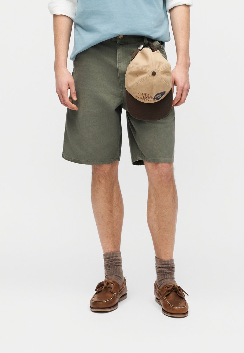 Homme portant un short vert olive, des chaussures bateau marron avec des chaussettes, tenant une casquette beige et marron à côté de lui.