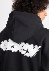 Black hoodie made of soft fabric with a white "obey" logo prominently displayed on the back. Features a large hood and relaxed fit.