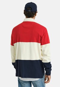 Lange mouwen pullover in rood, wit en marineblauw; heeft een kleurblokkenontwerp met een zachte textuur en een relaxte pasvorm. Kraag en detailing aan de achterkant zijn zichtbaar.