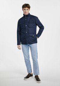 Granatowa, lekka kurtka z wysokim kołnierzem, dwoma kieszeniami na piersi, dwiema dolnymi kieszeniami i zapięciem na napy. W komplecie z jasnymi dżinsami i ciemnymi butami.