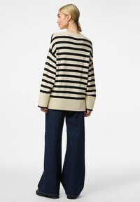 Sweter w paski w kolorze kremowym i czarnym, w oversize'owym kroju z ribbami na mankietach i dolnej części. Połączony z szerokimi niebieskimi dżinsami.