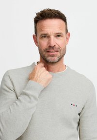Pull en texturé gris clair avec un col rond et des poignets côtelés. Présente un petit logo brodé en bleu, rouge et blanc sur la poitrine.