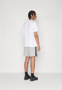 Λευκό μπλουζάκι, γκρίζα αθλητικά σορτς με μαύρες ρίγες, μαύρα παπούτσια. Λείο ύφασμα και χαλαρή εφαρμογή με απλό σχέδιο.