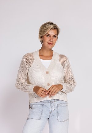 Frau mit kurzen blonden Haaren, die eine beige, geknöpfte Netzstrickjacke über einem weißen Top und hellblauen Jeans trägt, lächelnd mit verschränkten Händen.