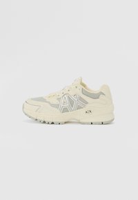 Chaussure de sport beige clair avec une tige en mesh, des accents argentés, un col rembourré et une semelle texturée. Présente le logo "AX" sur le côté.