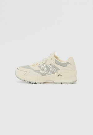 Zapatilla deportiva de color beige claro con parte superior de malla, acentos plateados, cuello acolchado y suela texturizada. Presenta el logo "AX" en el lateral.