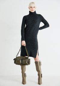 Charcoal gray turtleneck sweater dress with side slits, paired with a green shoulder bag featuring multiple pockets and beige knee-high boots.