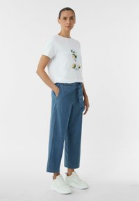 Witte t-shirt met een kleurrijke drankafbeelding, gecombineerd met losse blauwe denimbroeken en witte sneakers. Kenmerken zijn een ronde halslijn en zijzakken.