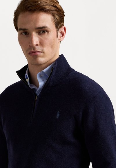 Marineblauwe gebreide pullover met een ritskraag, met een subtiel logo op de borst. Het materiaal lijkt zacht en gewafeld.