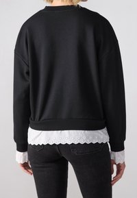 Sweat-shirt noir en tissu texturé, avec une bordure en dentelle à œillets blanche à l'ourlet et aux manches, associé à un jean foncé.