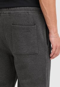 Gray cotton joggers with a textured finish, featuring an exterior back pocket, elastic waistband, and a close-up of a hand resting nearby.