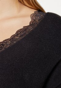 Zwarte sweater met een zachte textuur en een kantdetail langs de halslijn. Het kant heeft een bloemenpatroon, wat een delicate accent toevoegt.