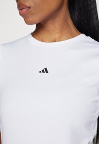 Närbild av en person som bär en vit träningsskjorta med en liten svart Adidas-logotyp p å bröstet, som visar hals och läppar.