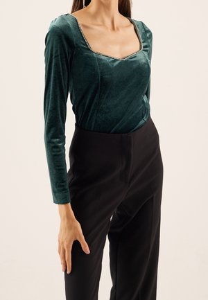 Kvinde iført en grøn velour langærmet top med firkantet halsudskæring og sorte højtaljede bukser mod en ensfarvet baggrund.