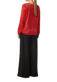 Rote, durchsichtige Bluse mit langen Ärmeln und rundem Ausschnitt, kombiniert mit lockeren schwarzen Hosen. Glatte Textur und fließendes Design.