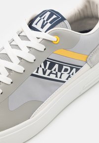 Sneaker grigia con tomaia liscia in pelle e mesh, con una striscia gialla, lacci bianchi e un logo stampato sul lato.