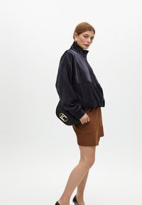 Kvinna i en marinblå zip-up tröja och bruna mockashorts, bärande en liten svart väska med en gyllene logotyp, står mot en enkel vit bakgrund.