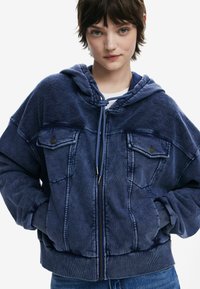 Blaue Kapuzenjacke mit Reißverschluss, zwei vorderen Taschen, gerippten Bündchen und Saum, tonalem Stoffmuster und lässiger Passform.