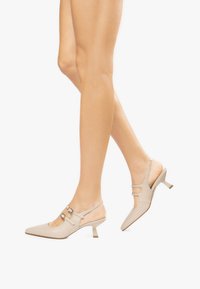 Tacchi slingback in pelle beige con punta affusolata, tacco basso a blocco e due cinturini alla caviglia regolabili. Texture liscia, design minimalista.