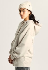 Junge Person mit dunklen Haaren und Nasenring trägt eine cremefarbene Mütze und einen übergroßen cremefarbenen Hoodie, steht seitlich vor einem schlichten, hellen Hintergrund.