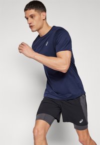 Marineblaues Sport-T-Shirt mit kurzen Ärmeln, kombiniert mit schwarz-grauen Shorts, entworfen für aktive Bewegungen. Mit Logo auf der Brust.