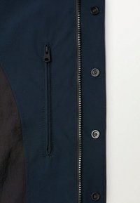 Námořnicky modrá bunda s hladkou texturou, vybavená předním zipem a patentními knoflíky. Na levé straně kapesní kapsa na zip.