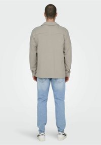 Beige, long-sleeve jacket with a shirt collar, smooth texture, and back yoke detail, paired with light blue denim jeans and white sneakers.