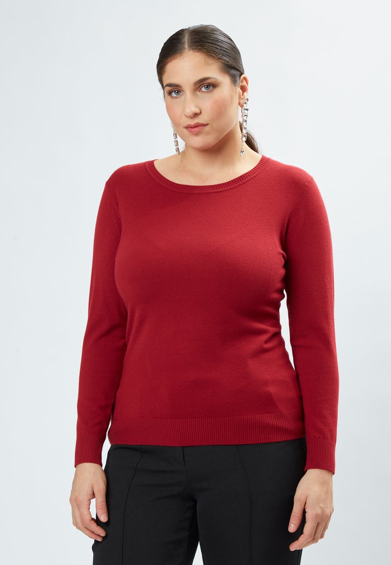 Roter Pullover mit langen Ärmeln, geripptem Halsausschnitt und Saum, aus weichem Material. Glatte Textur, taillierte Passform, kombiniert mit schwarzen geraden Hosen.