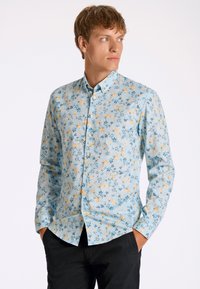 Ljust blå skjorta med långa ärmar som har ett blommönster med gula och blå blommor, vita knappar och en klassisk kragsdesign.