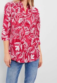 Femme portant une chemise à manches longues fleurs rouges avec des poignets retroussés et un jean bleu, se tenant devant un fond clair.