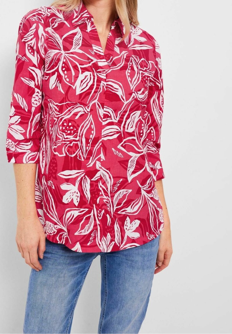 Femme portant une chemise à manches longues fleurs rouges avec des poignets retroussés et un jean bleu, se tenant devant un fond clair.