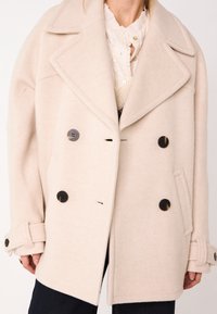 Cappotto di lana beige doppiopetto con grandi rever a punta, bottoni neri e tasche laterali; presenta una vestibilità rilassata e un tessuto testurizzato.