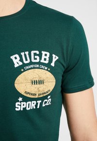 Camiseta de algodón verde con un diseño gráfico de balón de rugby y texto en blanco y dorado. Manga corta y diseño de cuello redondo.