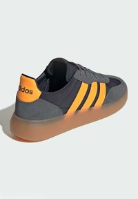Black and gray sneaker with orange accents, featuring three stripes, a suede upper, and a translucent rubber sole.