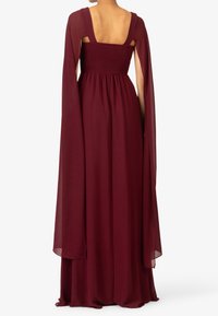 Nainen, joka käyttää lattiaan ulottuvaa, ilman hihoja olevaa burgundin punaista mekkoa, jossa on leveät olkaimet ja kiinnitetyt läpinäkyvät viittahihat, näkyy selkäpuolelta.