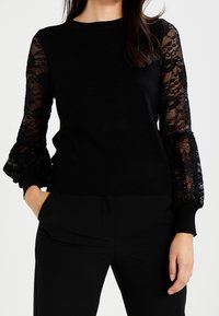Pull noir avec des manches longues en dentelle, des poignets et un ourlet côtelés. Le corps est en tricot noir uni. Design facile à porter ; minimalisme dans les détails.