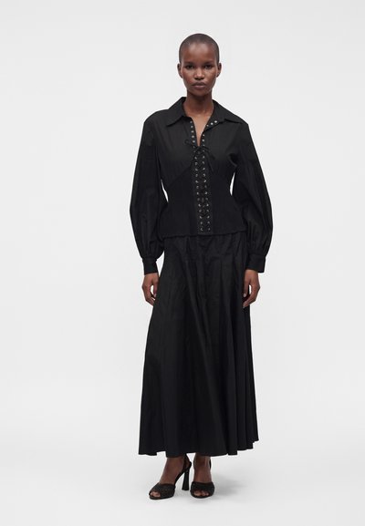 Femme portant un chemisier noir à manches longues avec laçage à l’avant et une longue jupe noire, debout devant un fond blanc uni.