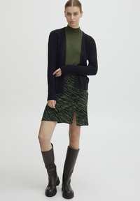 Svart ribbad cardigan över en grön polotröja, matchad med en grön kjol i zebra-mönster med knappdetaljer och en slits på sidan, kompletterad med bruna knähöga stövlar.