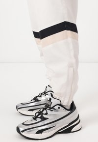 Pantalones deportivos blancos con rayas horizontales en negro y beige, combinados con zapatillas deportivas plateadas con acentos negros y superficies texturizadas.