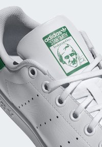 Baskets blanches en cuir avec une semelle plate, présentant une touche de vert au talon et une étiquette portant le nom "Stan Smith". Les lacets sont blancs.