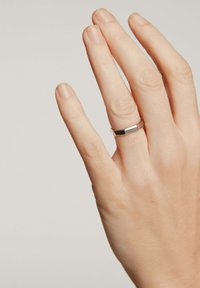 Anillo de plata con una parte superior rectangular plana, superficie lisa y acabado pulido, llevado en una mano con uñas naturales sobre un fondo neutro.