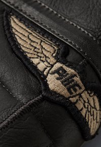 Material de cuero negro con un destacado logotipo bordado de alas en beige, que presenta un detallado cosido y una superficie texturizada.