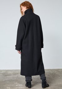 Lang sort uldfrakke med høj krave, vide ærmer og knapdetaljer ved manchetterne. Glat tekstur med et minimalistisk design.