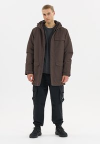 Braune isolierte Jacke mit Kapuze, Frontreißverschluss und mehreren Taschen. Kombiniert mit schwarzen Cargo-Hosen und dunklem Schuhwerk.