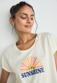 Cream-colored short-sleeve T-shirt featuring a colorful sunburst graphic with the text "Hey Sunshine" in navy blue. Soft fabric and relaxed fit.