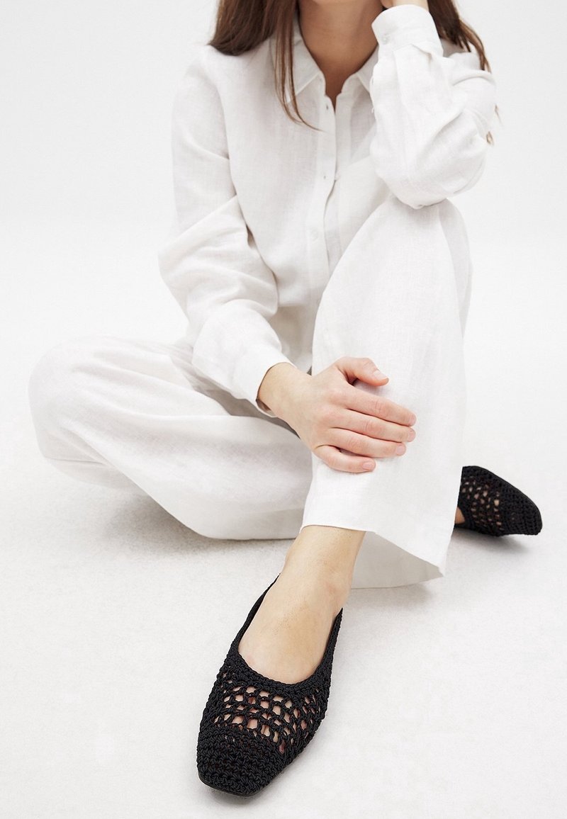 Chaussures plates noires en crochet avec un design en maille ouverte et un bout solide, associées à un pantalon ample en lin blanc et une chemise à boutons.