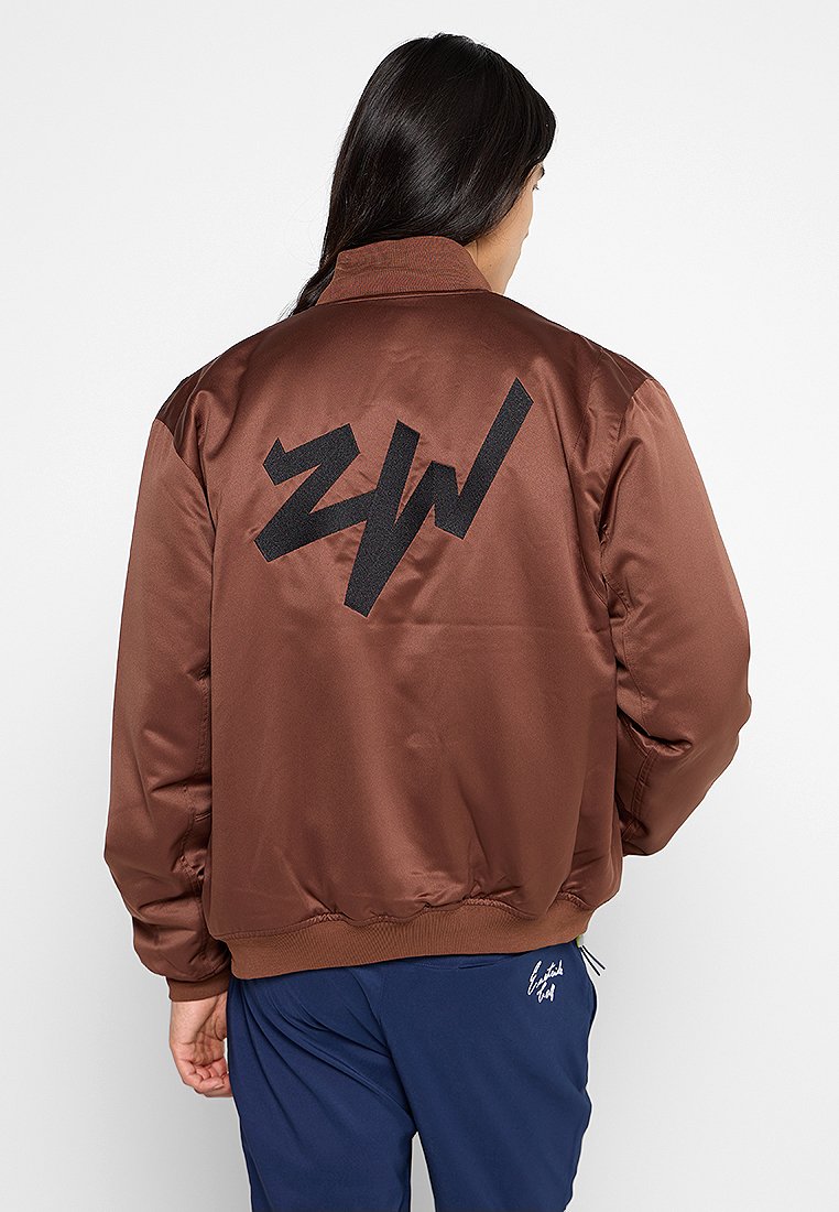 Giubbotto bomber in satin marrone con polsini e orlo a coste. Presenta un grande logo nero sul retro. Tessuto liscio, vestibilità rilassata.