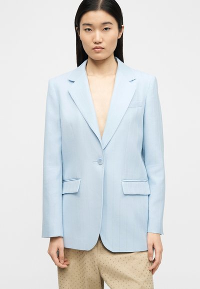 Γυναίκα που φοράει γαλάζιο σακάκι με μονό κουμπί και μπεζ παντελόνι με μικρές λεπτομέρειες eyelet, στέκεται μπροστά σε απλό φόντο.